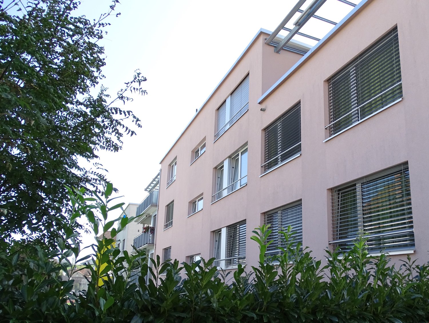 Multiple story building, pink paint, multiple windows with blinds, balcony, greenery