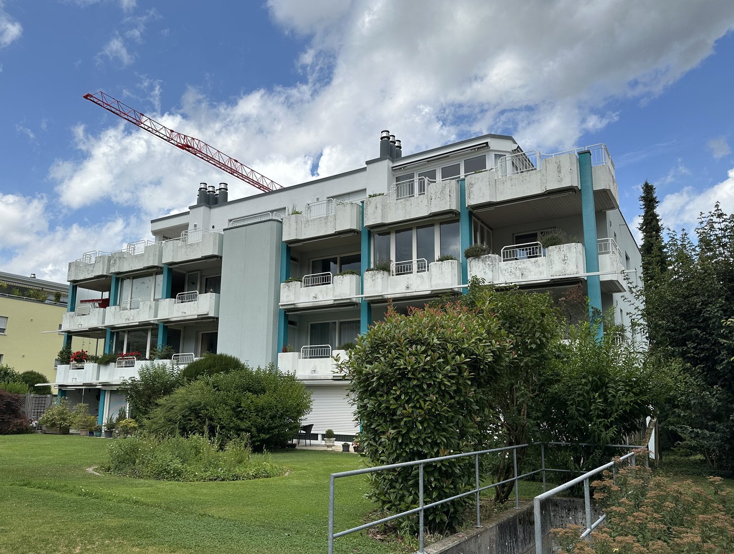 Multi-story apartment building with balconies, surrounded by greenery and a crane visible in the background