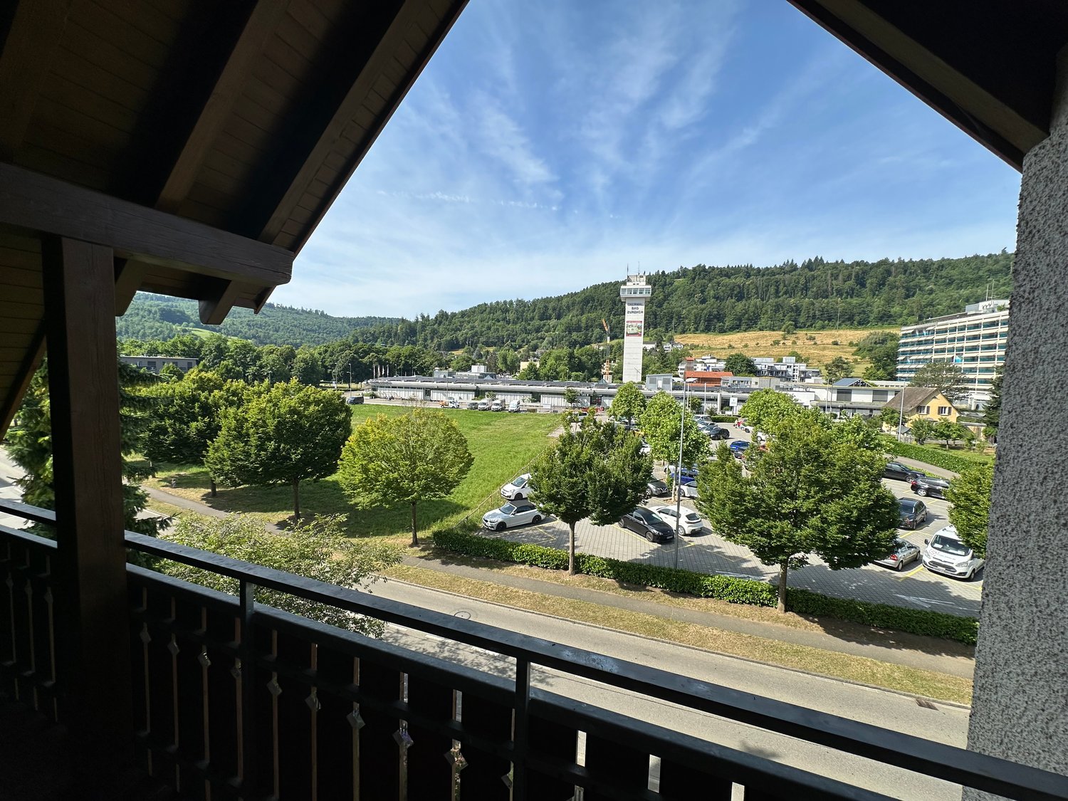 The image shows a view from a balcony or terrace overlooking a town or city. The scene includes a mix of buildings, trees, and a parking lot. The sky is blue with some clouds, and the surrounding hills and forests are visible in the distance.