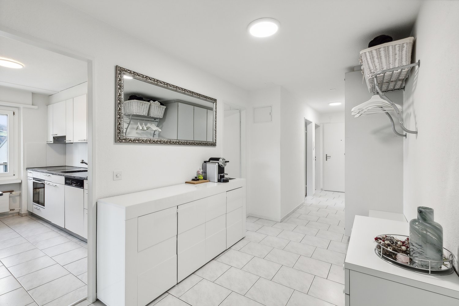 Modern white kitchen with ceramic tiles, white cabinets, kitchen counter, gas stove, sink, dishwasher, ceramic baskets, basket on the wall, hanging rack, lamp, mirror, table with a gray vase and a plate