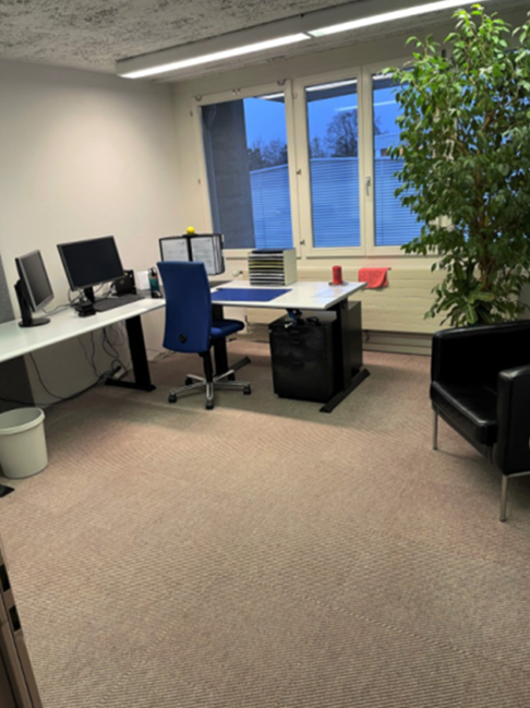 The image shows an office space with a desk, computer, and other office equipment. There is a large window with a view of what appears to be a balcony or terrace outside.