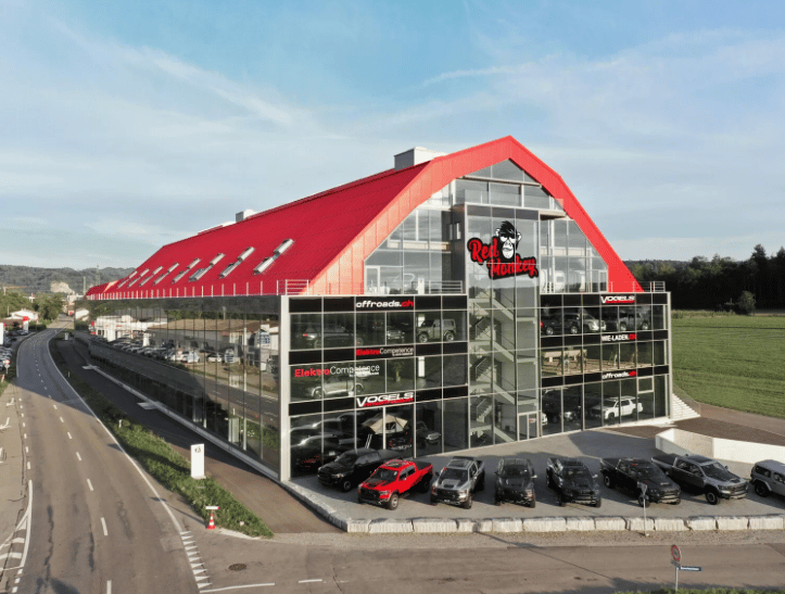 Modern building with red roof, glass walls, multiple cars parked in front, logo on the side