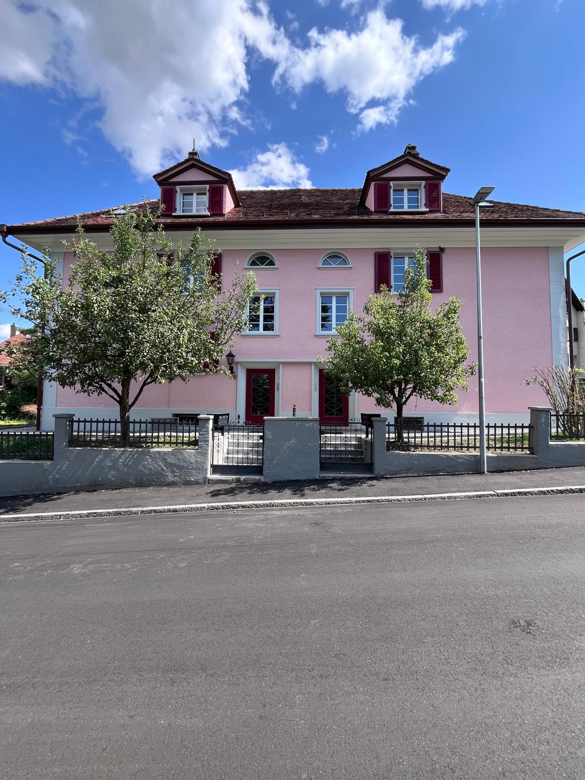 2-story pink building with red-tiled roof, arched windows, and balconies on the upper floors. The building is surrounded by trees and has a metal fence in front.