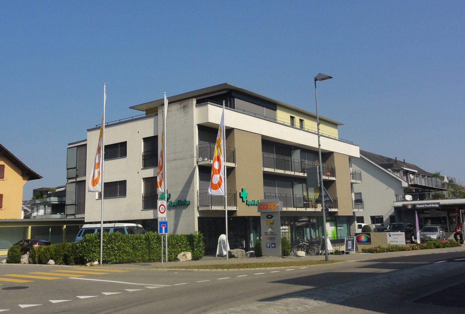 multi-story building with ground floor, pharmacy on ground floor, cars parked in front, greenery, streetlights, bicycles, bicycles