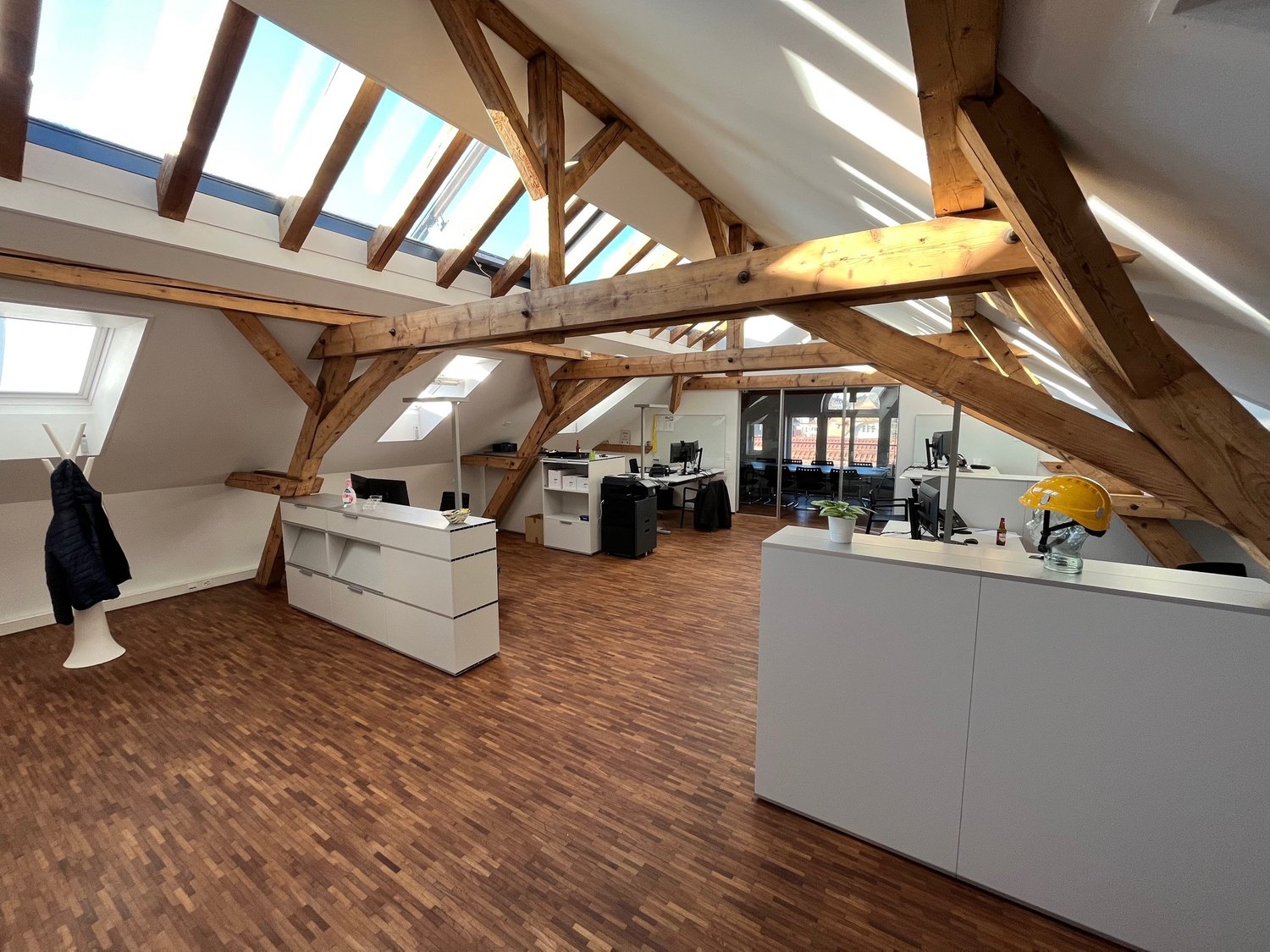 wooden beams, wooden floor, white walls, office desks, monitors, lamps, potted plants