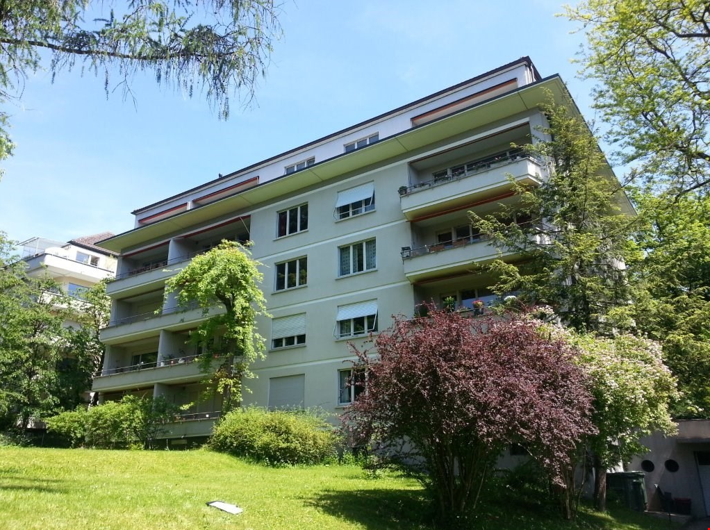 multi-storey residential building, white exterior, several balconies with railings, set in a park-like environment with lush greenery