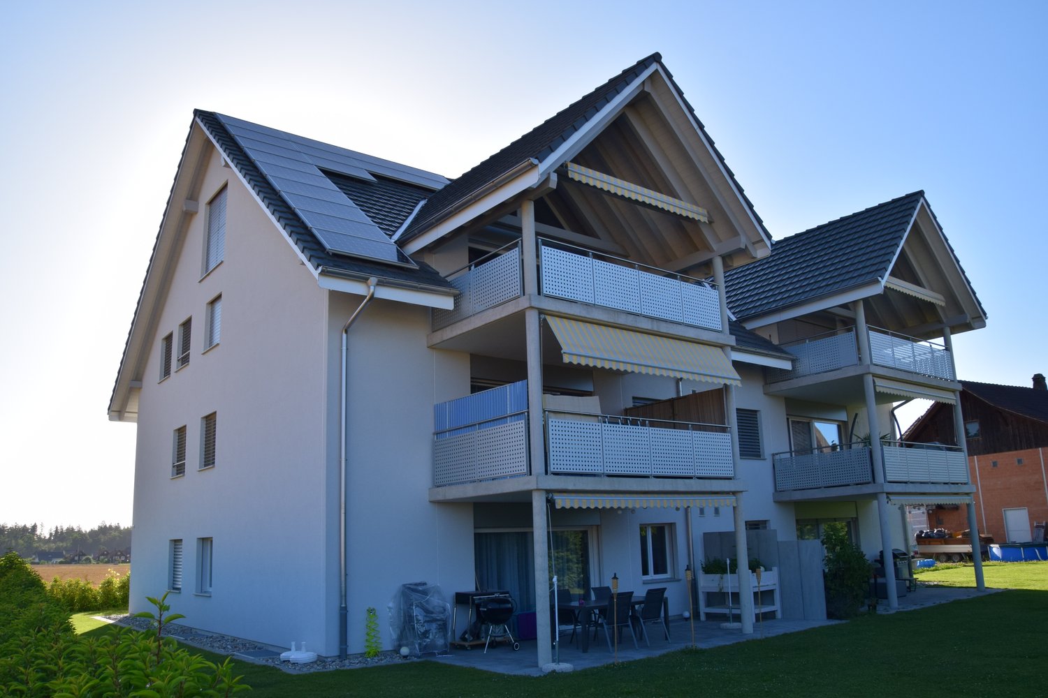 white building, 3 floors, balconies, solar panels, outdoor grill and table, grassy lawn, another house in the background