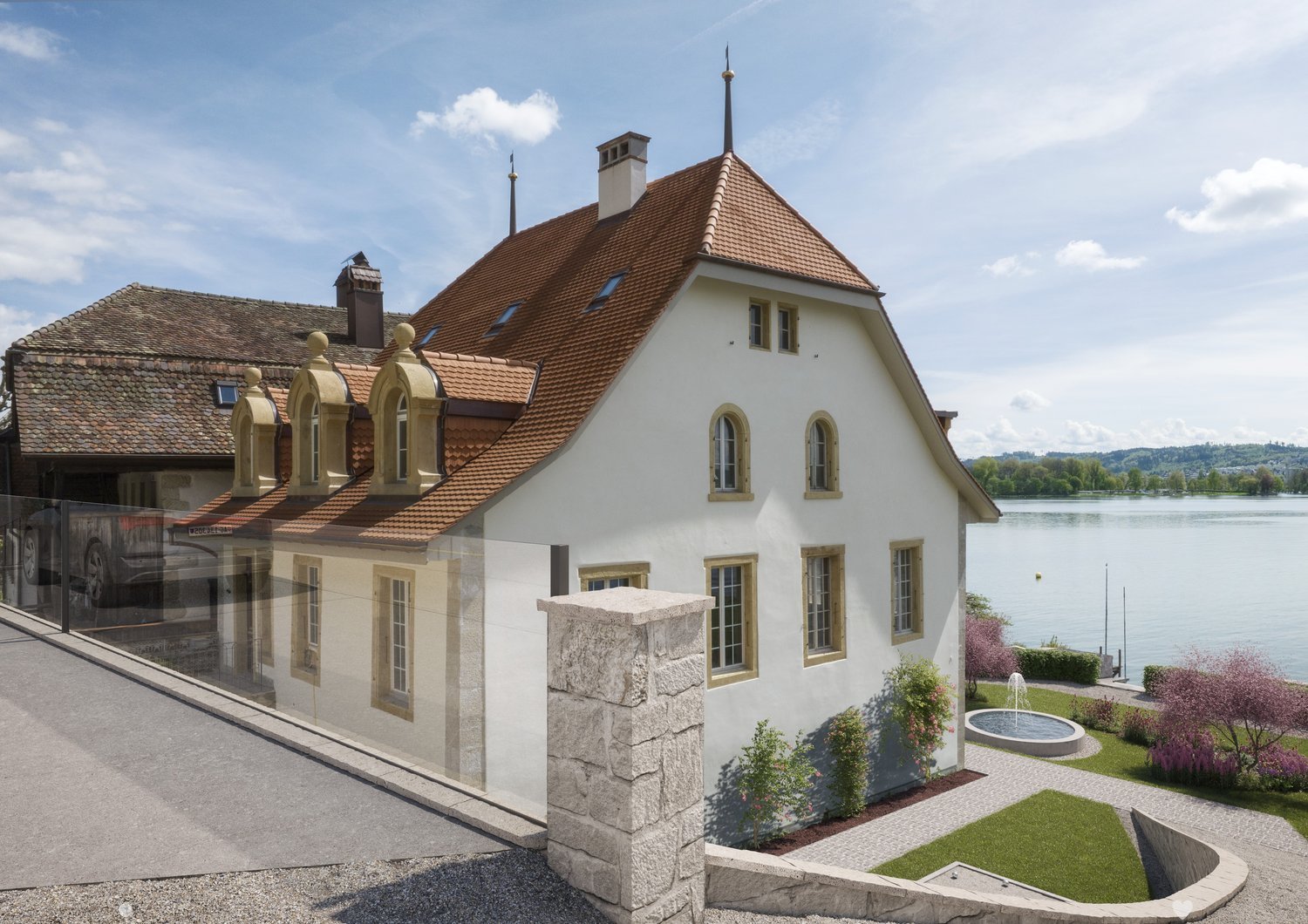 white house, brown roof, 3 dormers, multiple windows, located on lake, garden, small fountain