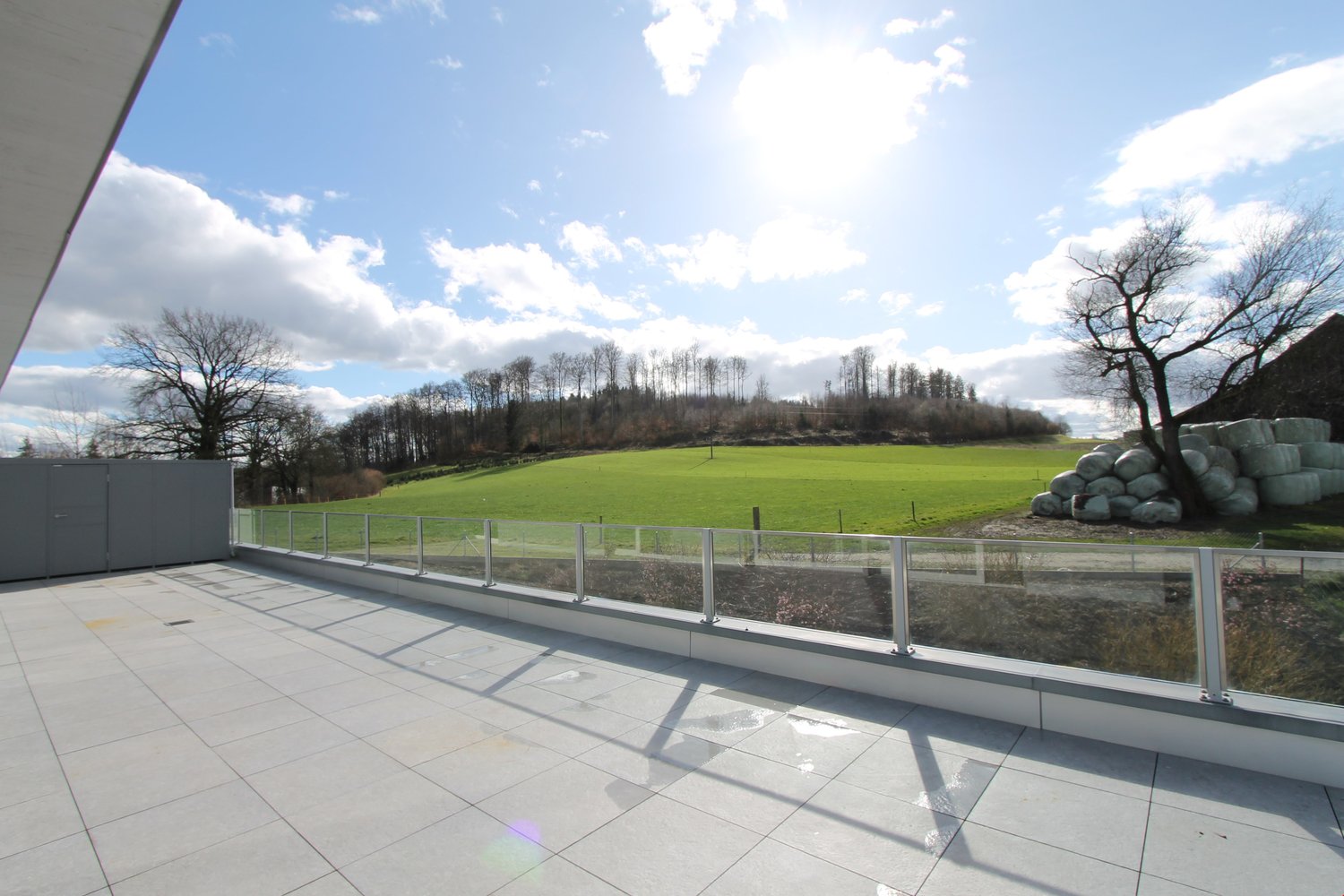 The image shows a large outdoor terrace or balcony with a glass railing overlooking a grassy field surrounded by trees. The sky is partly cloudy with some blue patches. The terrace appears to be part of a larger building or property.