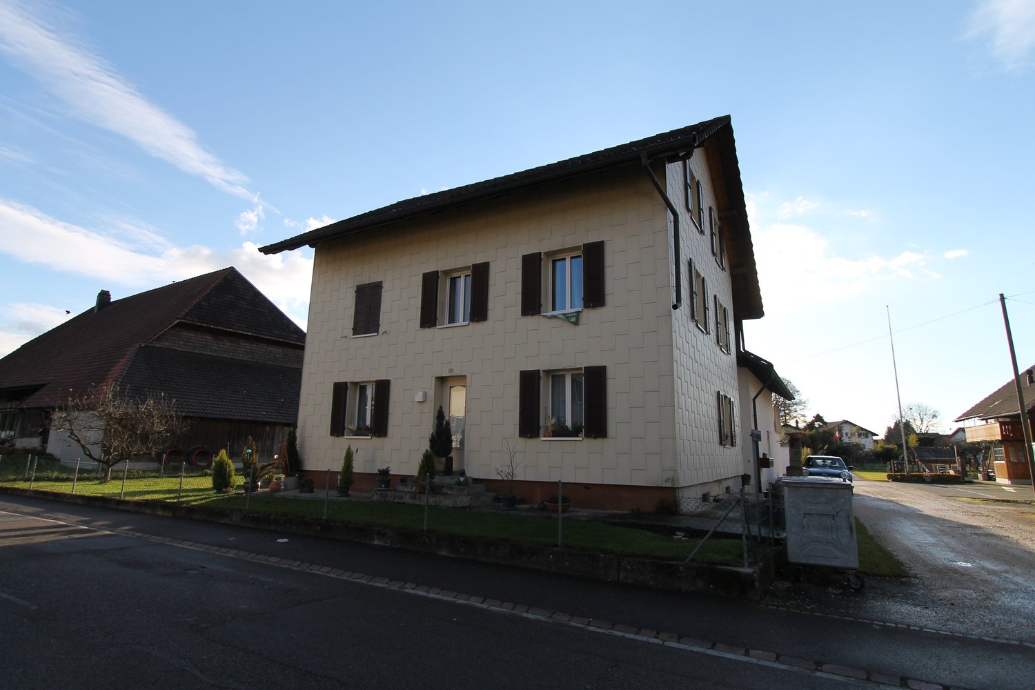 2-story residential building with white exterior walls, black shutters, and a slanted roof. There is a small garden area in front of the building and a parking space visible on the street.