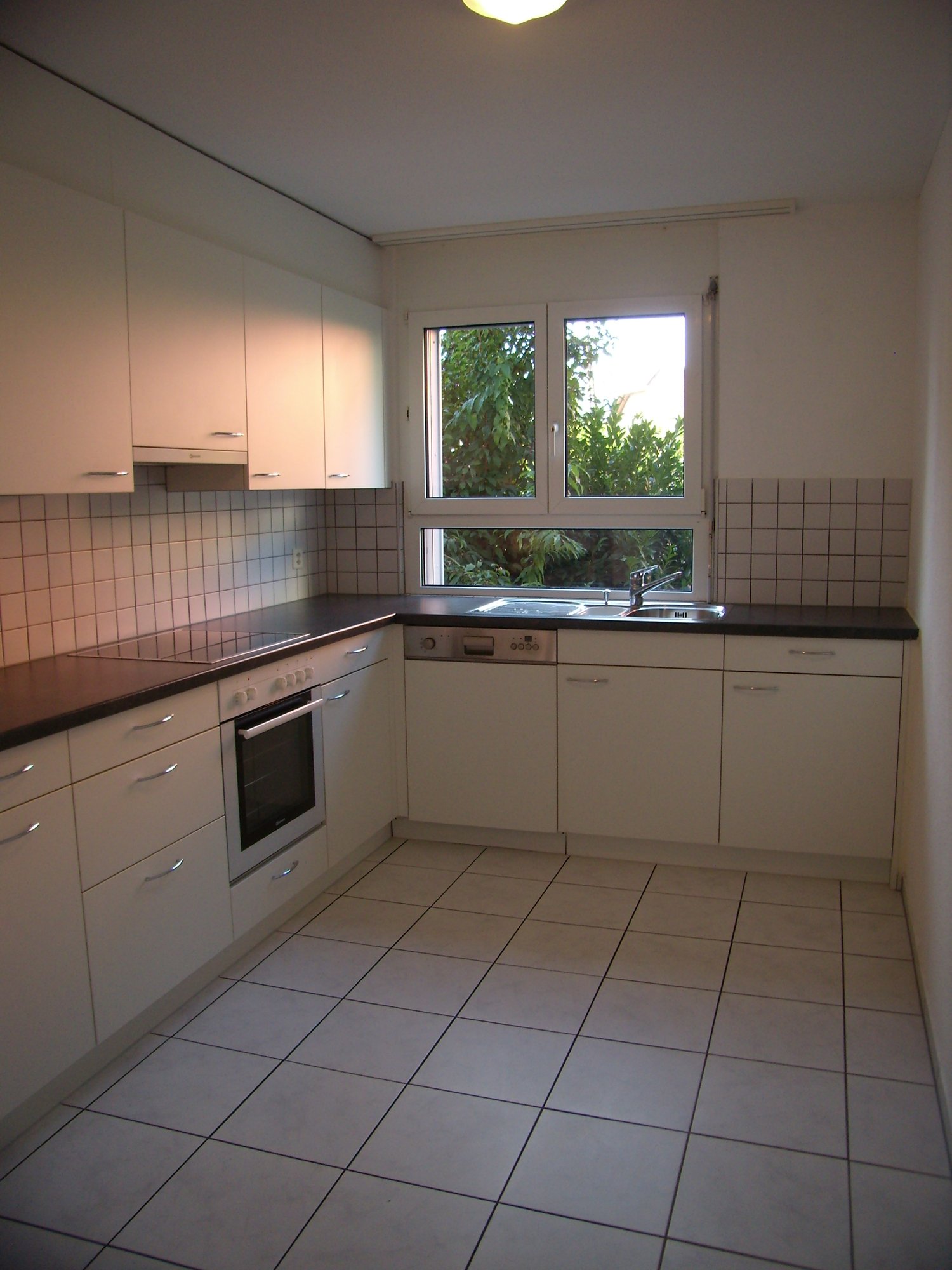 Fully fitted kitchen with tiled floor, white cupboards, gas hob, electric oven, dishwasher, sink with faucet, large window