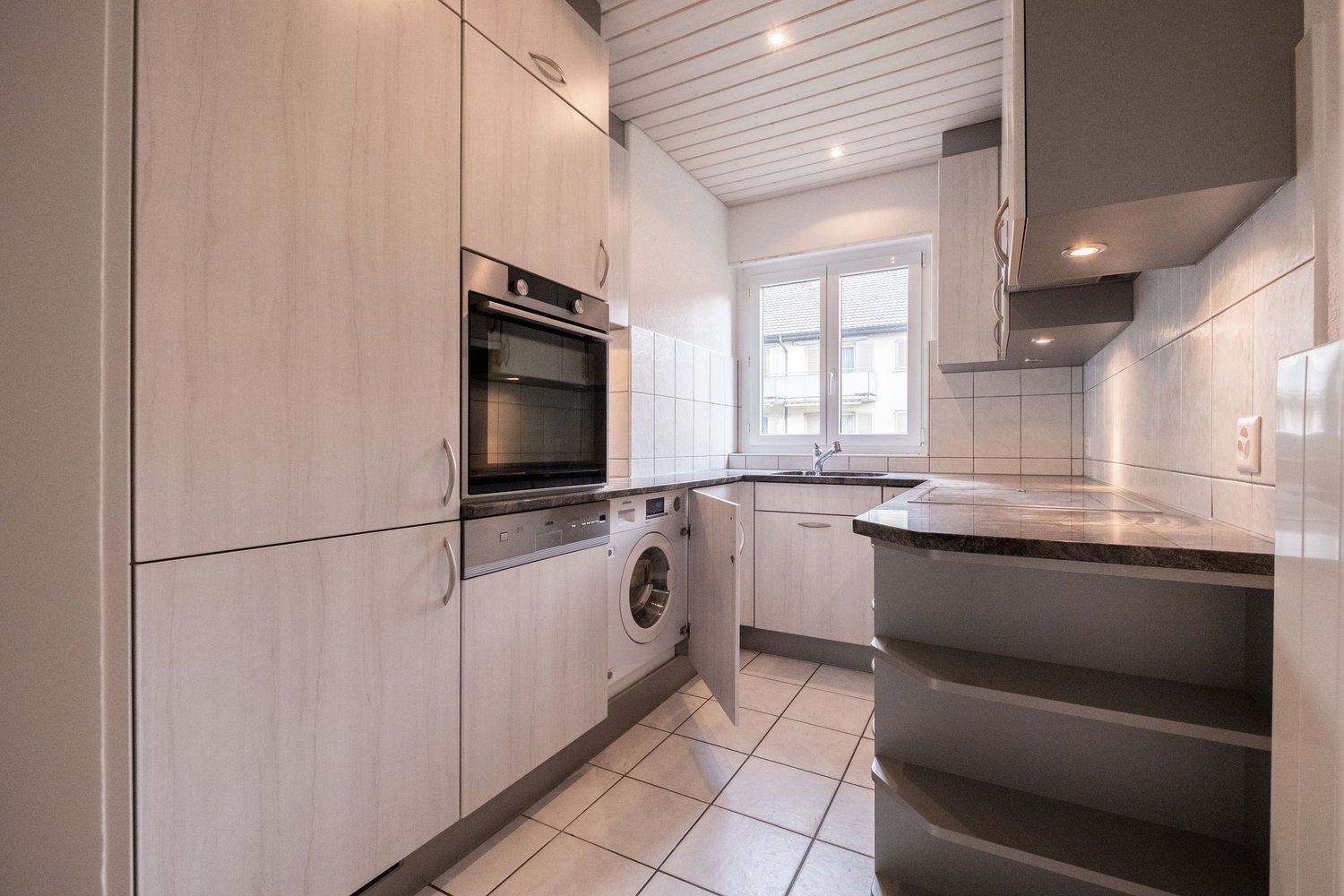 Stainless steel oven, white washing machine, white cupboards, tiled floor, sink with faucet