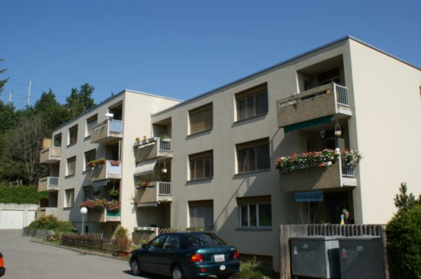 5-story white building with balconies, green plant boxes, a car parked in front, white garage on the side, trees in the background