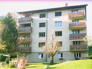 Multi-story apartment building with balconies, surrounded by trees and greenery