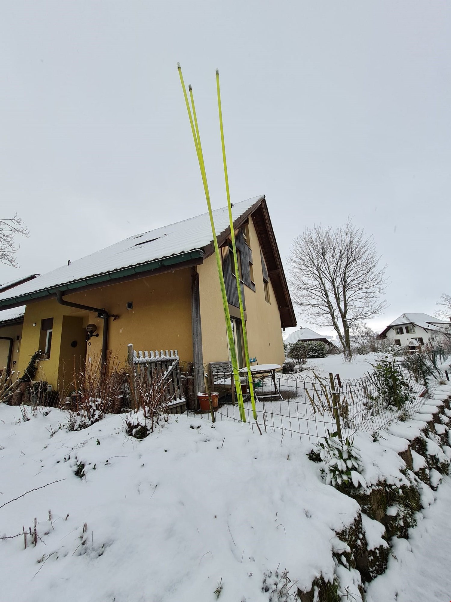 Yellow painted house, snow-covered rooftop, wooden fence around the house, yellow poles in the yard, snow covering the ground, trees with no leaves