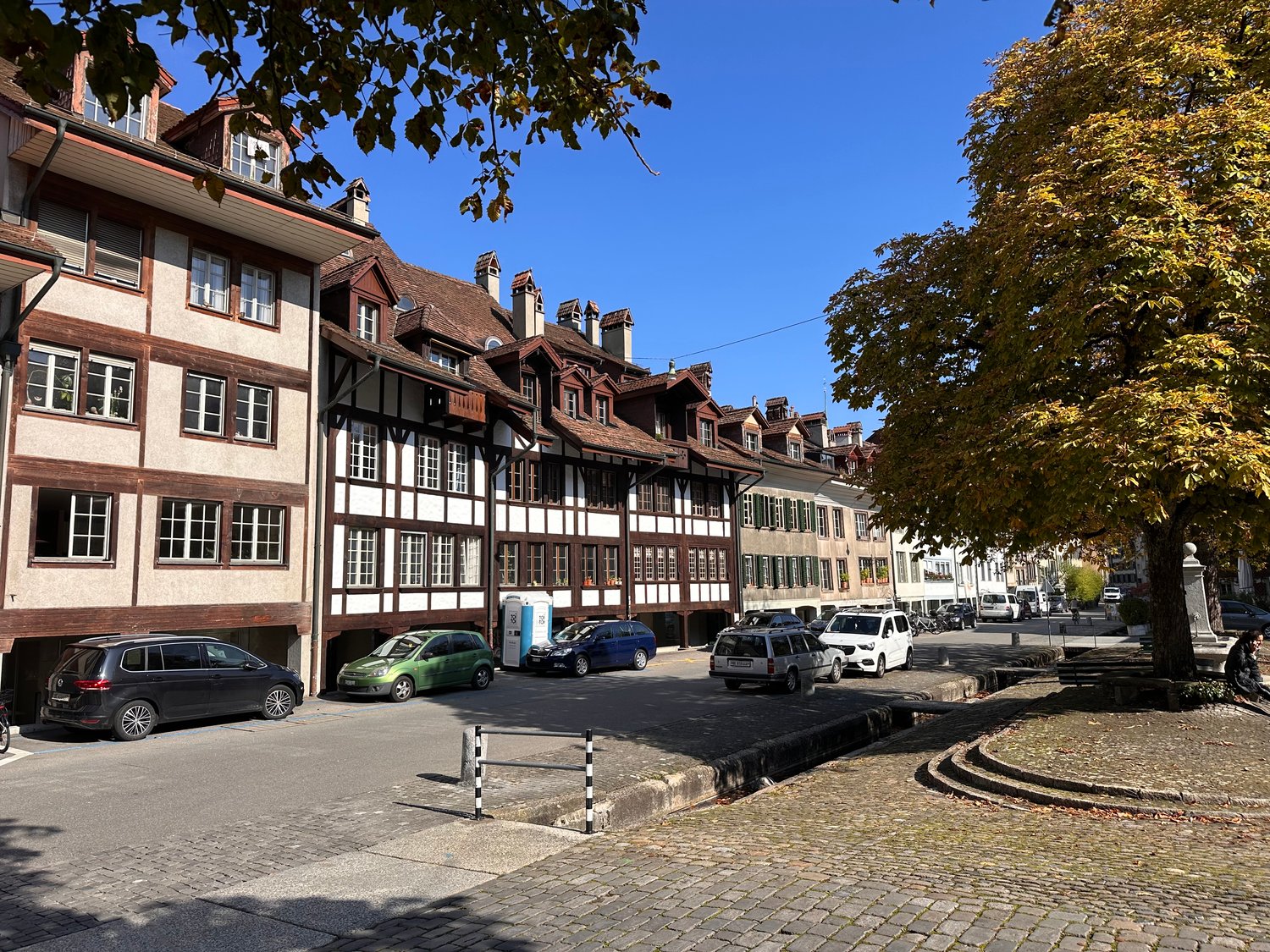 Multiple residential buildings along a street, with cars parked in front of some buildings. The buildings have brick and white exteriors, and a few are accessed through garages. Trees and utility poles are also visible.