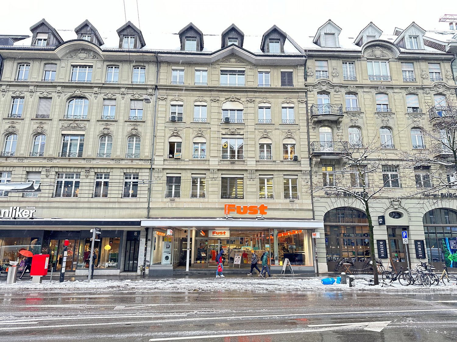 A multistory building with multiple entrances to stores, people walking in front, bicycles parked, trees, and a snowy ground