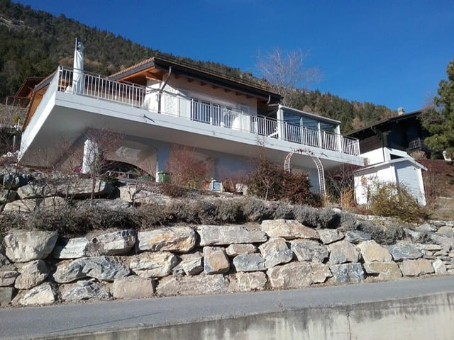 Two-story house, white exterior, balcony with railings, stone retaining wall, mountains in the background.