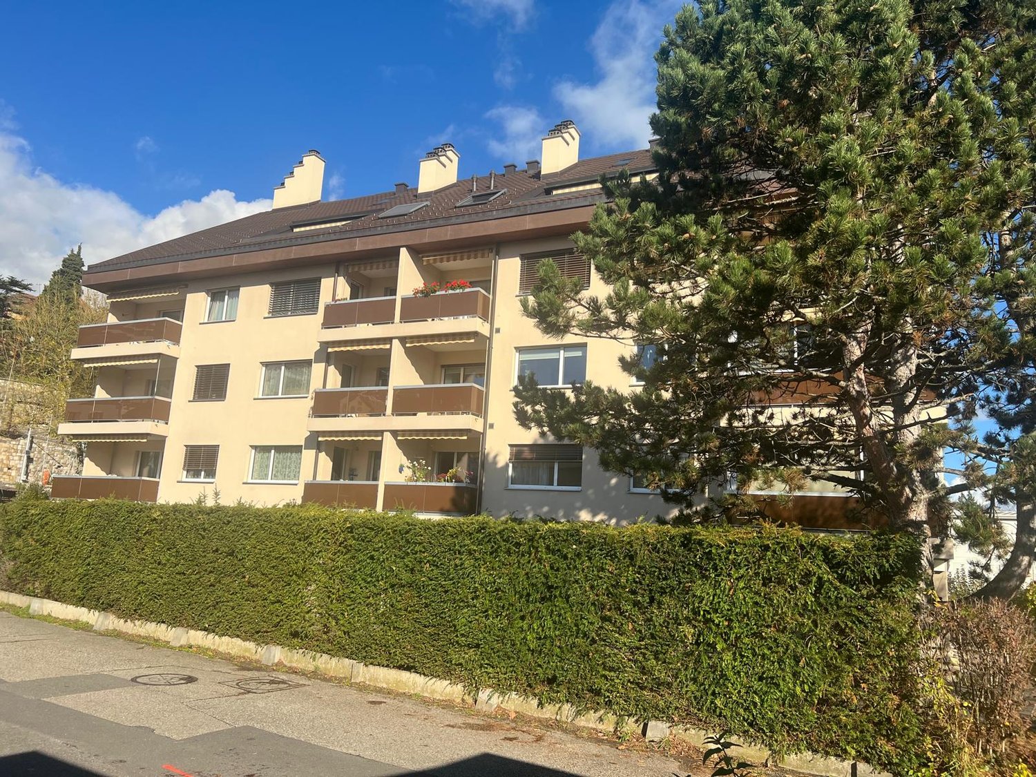 apartment building, beige facade, brown roof, balconies, windows with shutters, hedge, street
