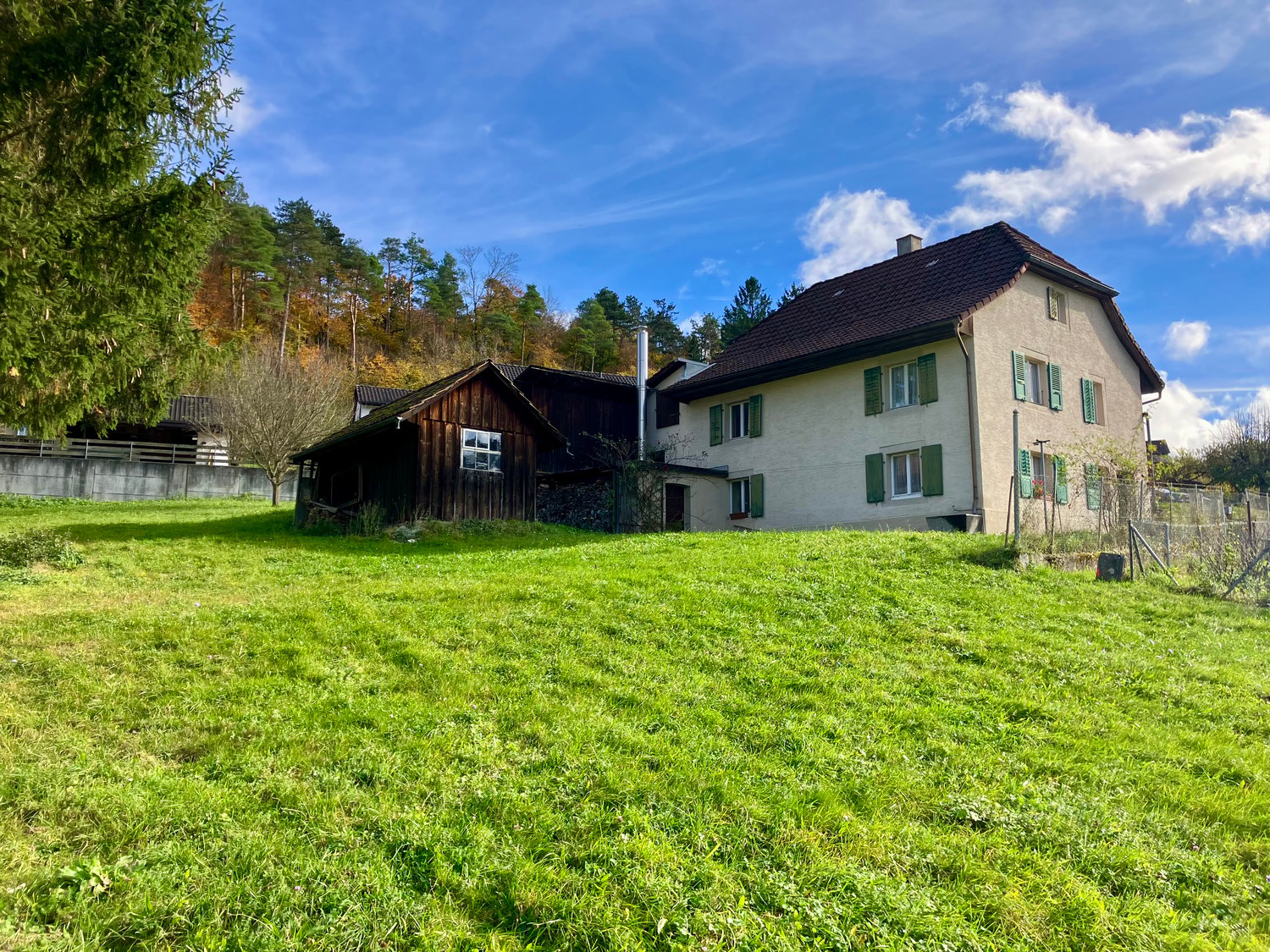 2 story house, brown roof, green shutters, wooden barn, large grassy field, trees in the background