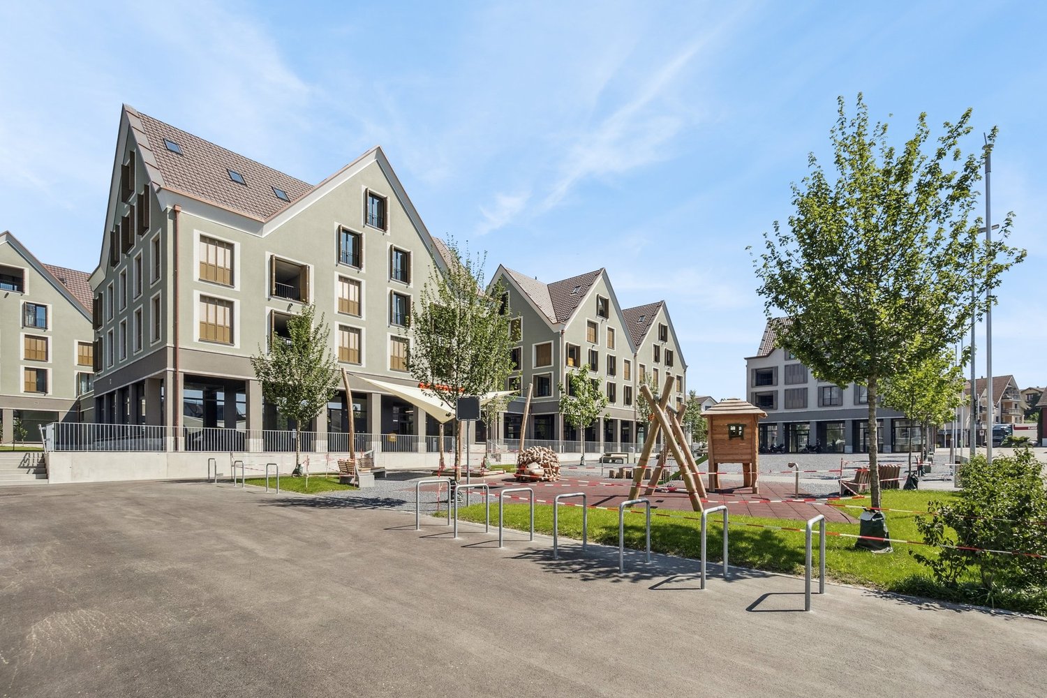 The image shows a residential area with several multi-story apartment buildings. The buildings have a modern, uniform architectural style with gabled roofs and large windows. There is a public plaza or square in the foreground with benches, bike racks, an
