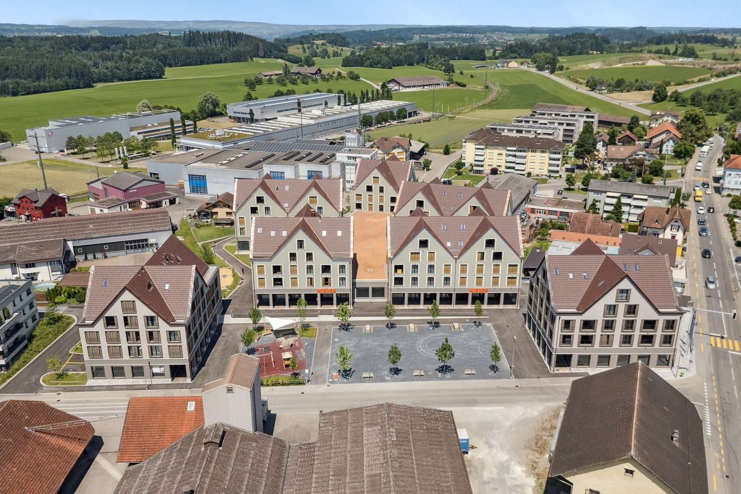 The image shows an aerial view of a town with a mix of residential and commercial buildings. The town has a central square or plaza surrounded by multi-story buildings with red tile roofs. There are also some industrial or warehouse buildings in the backg