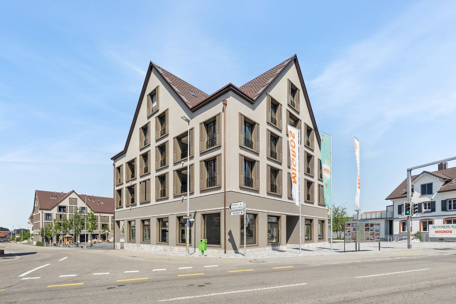 4-story commercial building with a modern, angular design. The building has a beige exterior with dark-colored roofing. There are multiple windows on each floor and a ground-level retail space. A parking area is visible in front of the building.