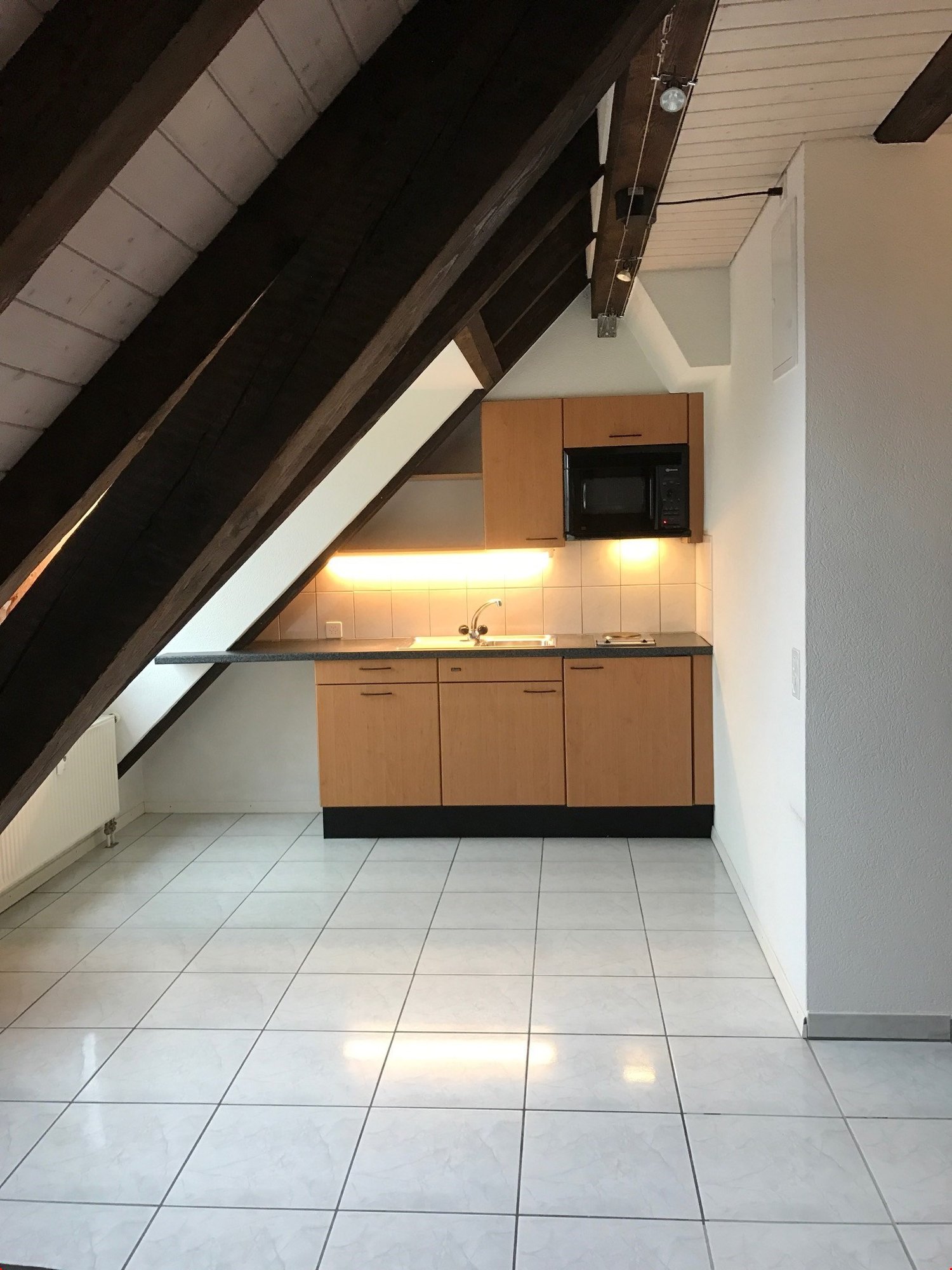 Small attic kitchen with wooden beams, tiled flooring, a countertop with a sink, and wooden cabinets. It includes a microwave and an electric stove.