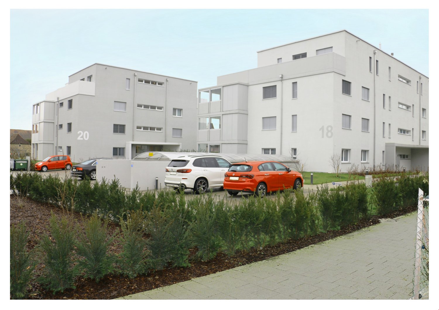 Two story white residential buildings, several parked cars, bushes