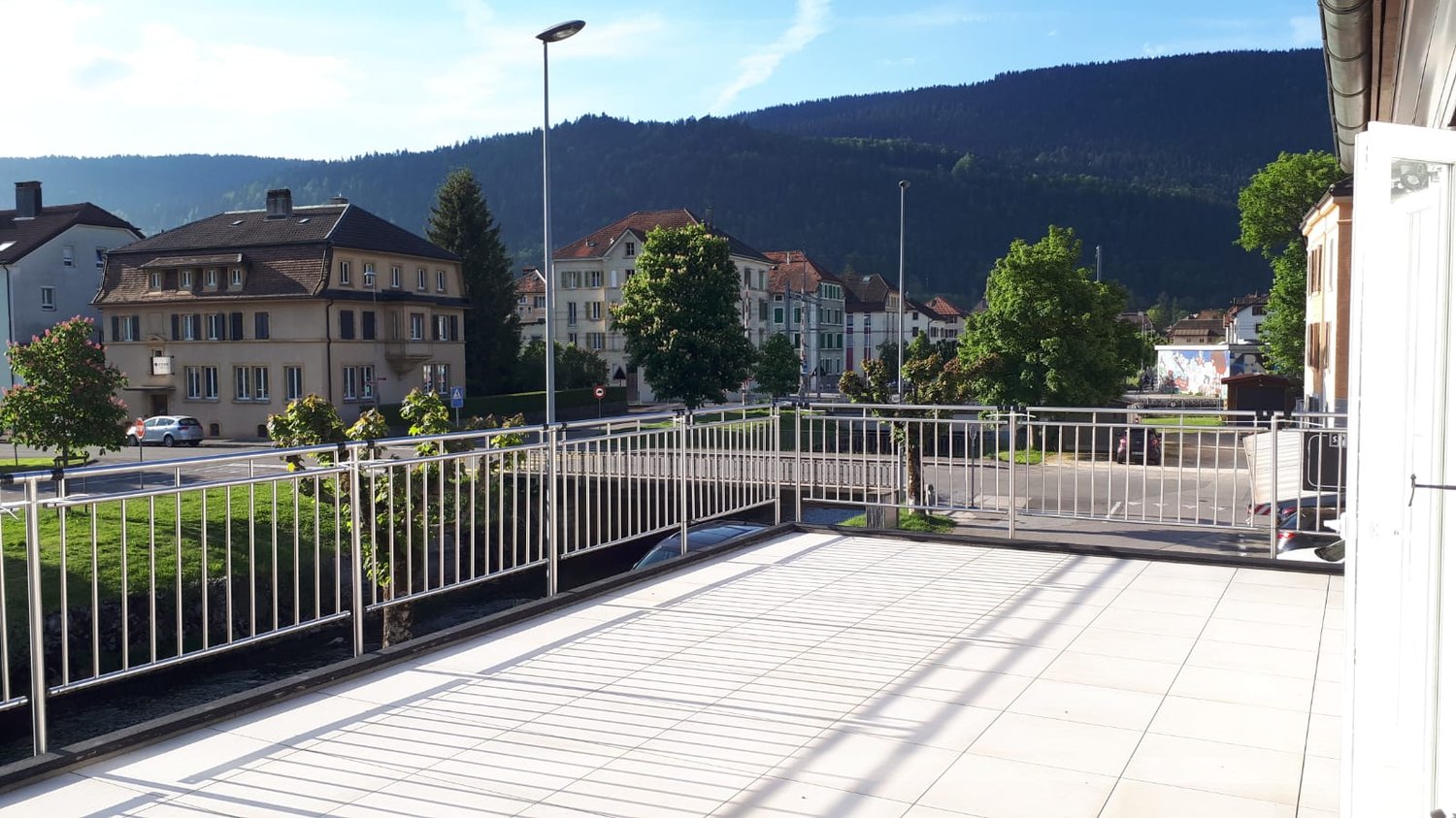 The image shows an outdoor area with a paved walkway and metal railings overlooking a town. There are several multi-story buildings in the background, surrounded by trees and mountains. The scene appears to be in a scenic, mountainous location.