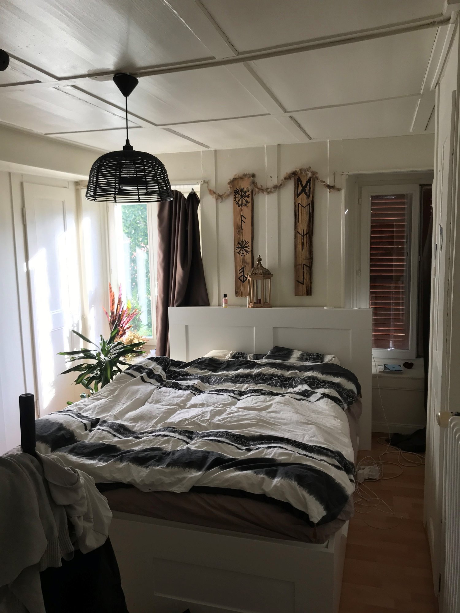 White bed, black and white striped blanket, bedside table with bottle, pendant lamp, white headboard, wooden floor, windows with brown curtains, plants, radiator