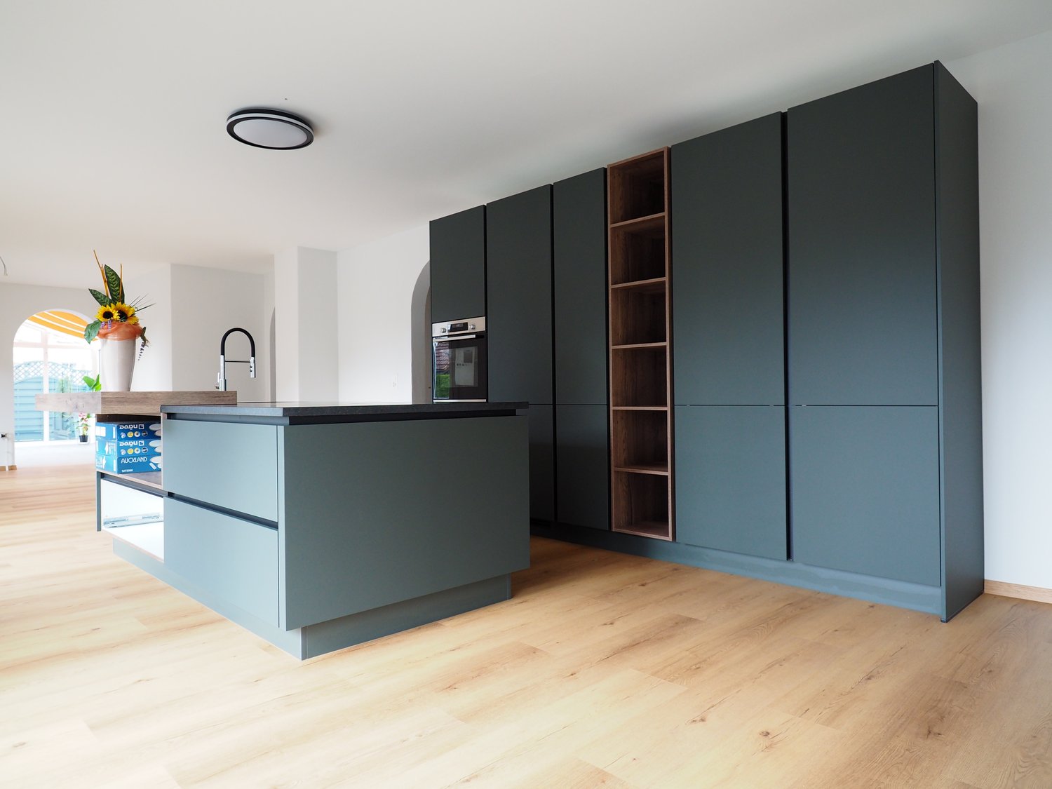 modern kitchen with a central island, dark grey cabinets, wooden countertops, integrated oven, built-in dishwasher, large sink, wooden shelves, flowers on countertop