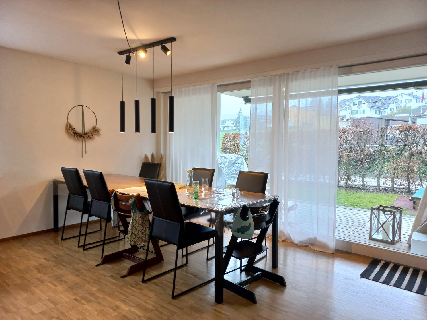 Dining room with wooden floor, long table, white curtains, black chairs, view of the outside through sliding doors.