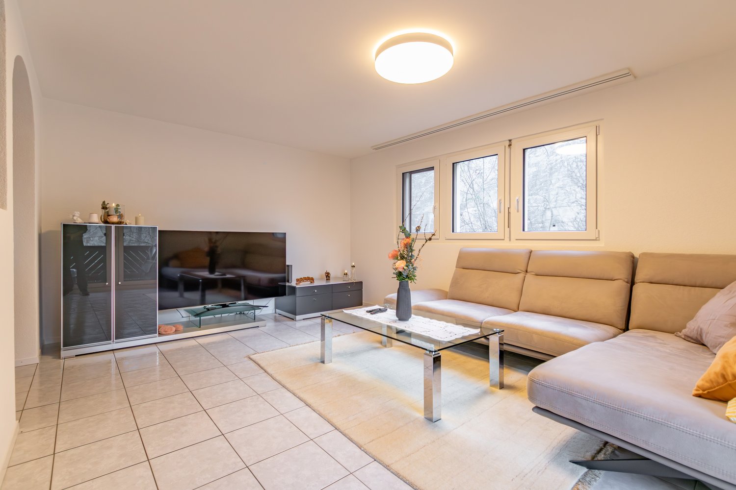 white tiles, beige sofa, glass coffee table, flat screen TV, cabinet, flowers, round lamp