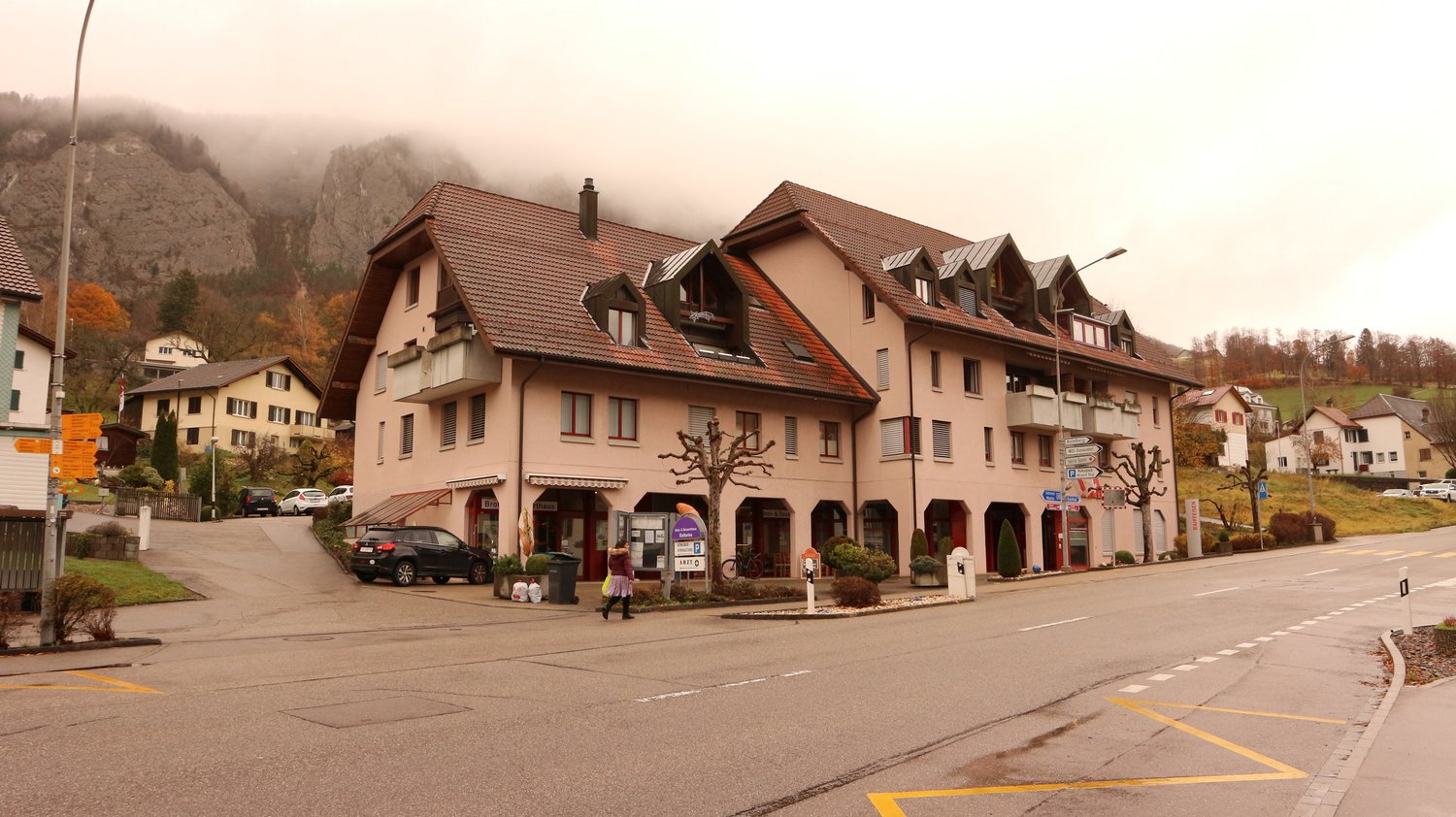 A row of multi-story residential buildings with red tile roofs, located in a mountainous area with fog in the background. The buildings have shops or businesses on the ground floor and apartments above. There are parked cars in front of the buildings.