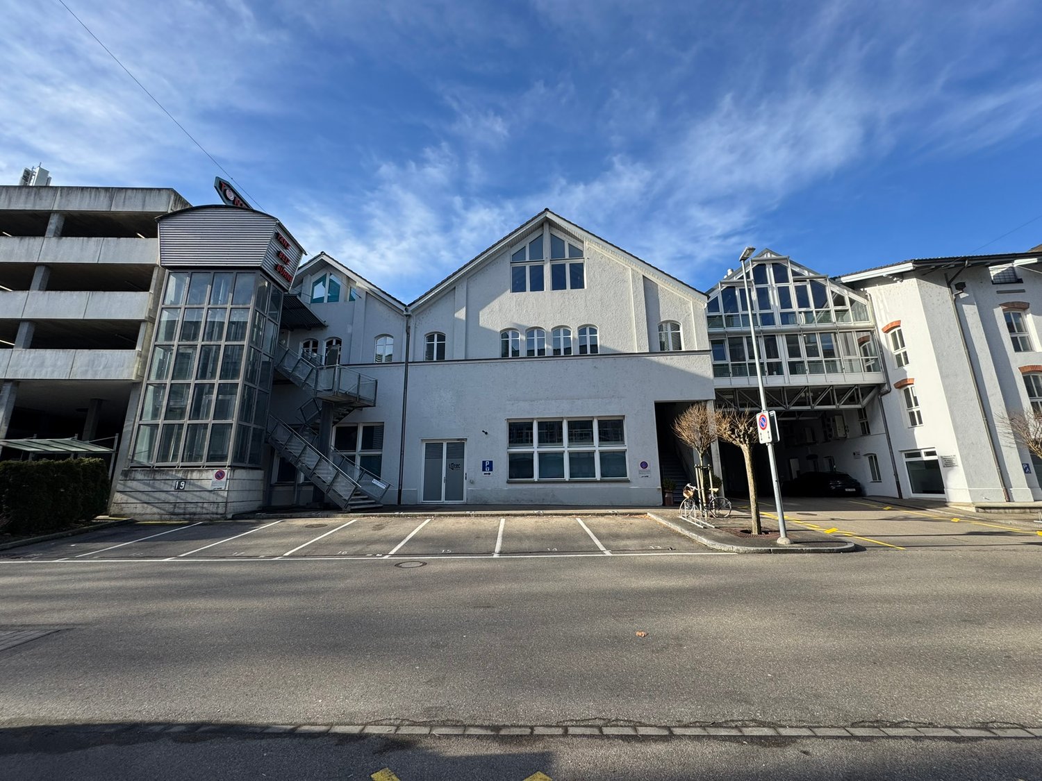 A multi-story commercial or office building with a modern, glass-heavy architectural design. The building has a large central structure with several wings and annexes. The exterior is primarily composed of light-colored walls and large windows. There is a