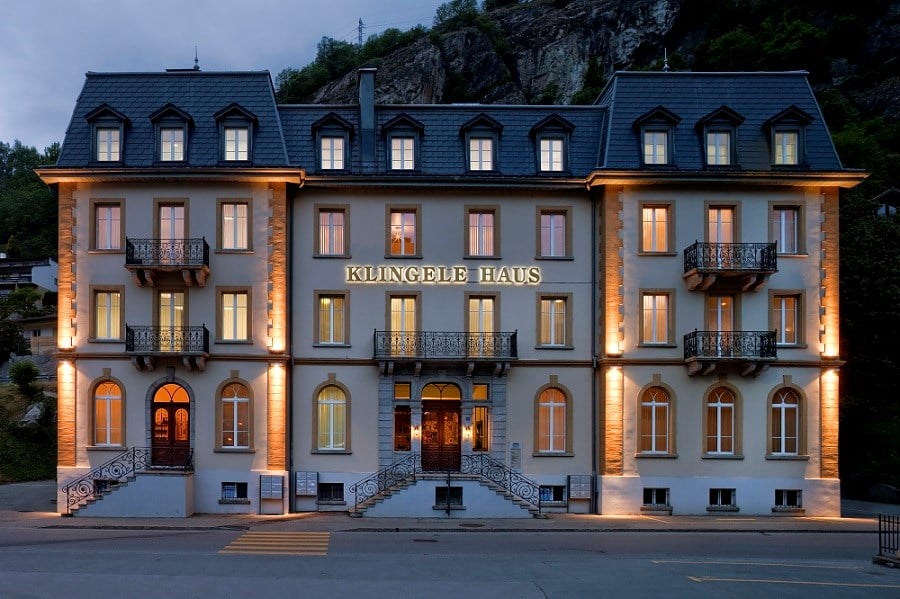 A multi-story hotel building with a traditional European architectural style, featuring stone walls, balconies, and large windows. The building is illuminated at night, creating a warm and inviting atmosphere. The name 'Klingele Haus' is prominently displ