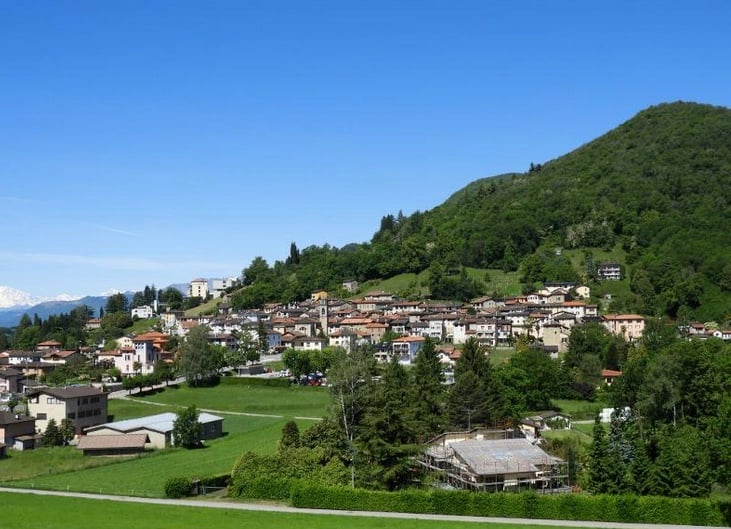 The image shows a picturesque mountain village nestled in a lush, green valley. The village is built on a raised ground, with houses and buildings scattered across the hillside. The landscape is dominated by the surrounding mountains, which are covered in