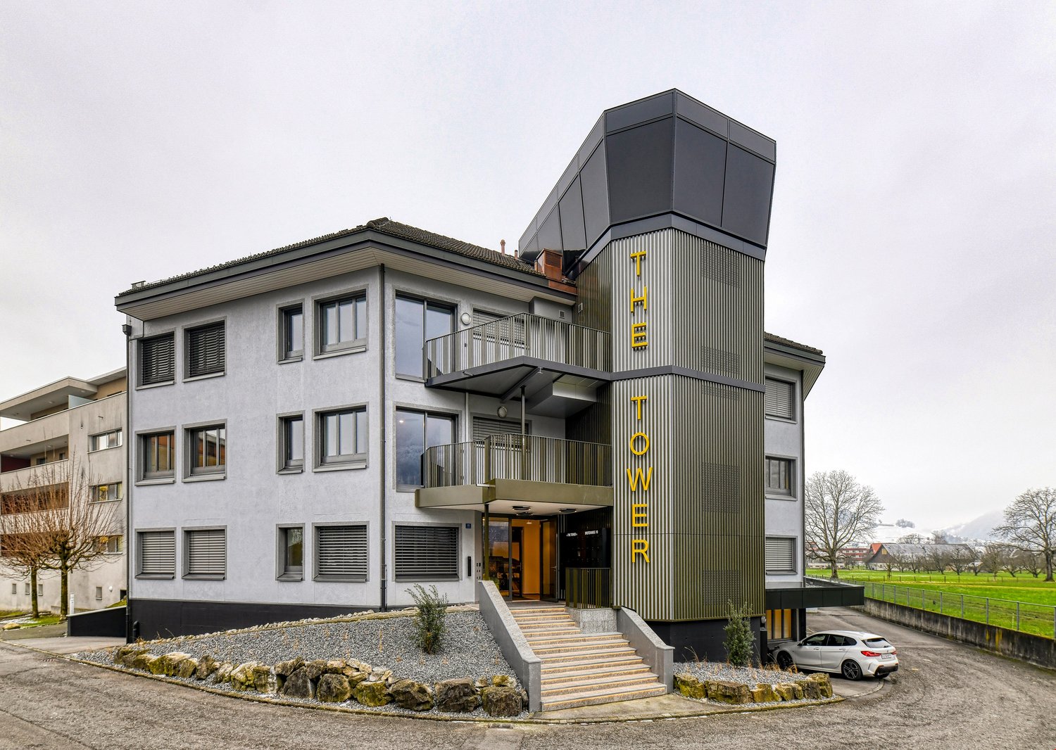 Contemporary building with multiple floors, modern facade, staircases leading to the entrance, car parked in front, and a surrounding landscape.