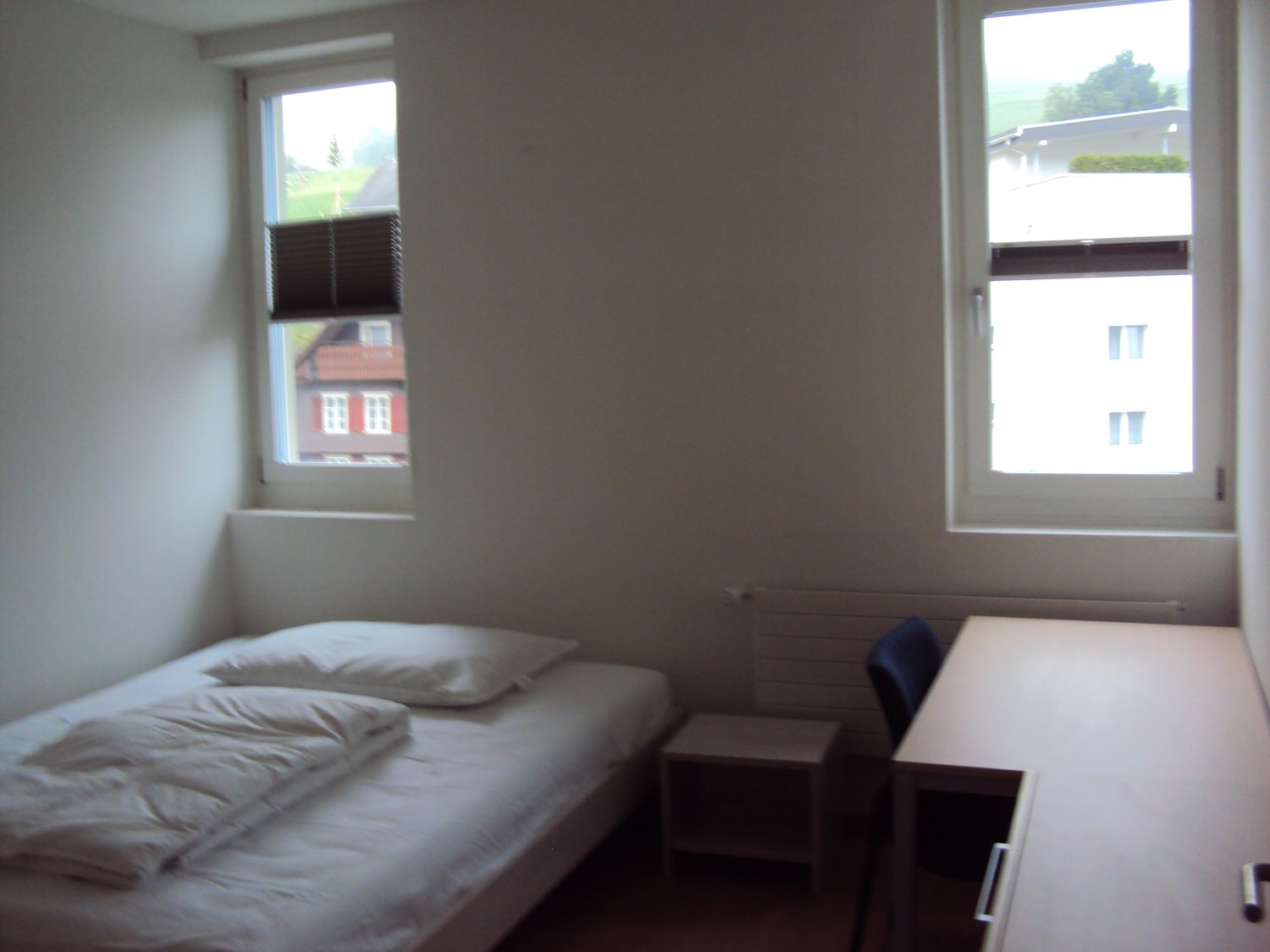 Single bed with 2 white pillows and a blanket, white desk and chair, white radiator
