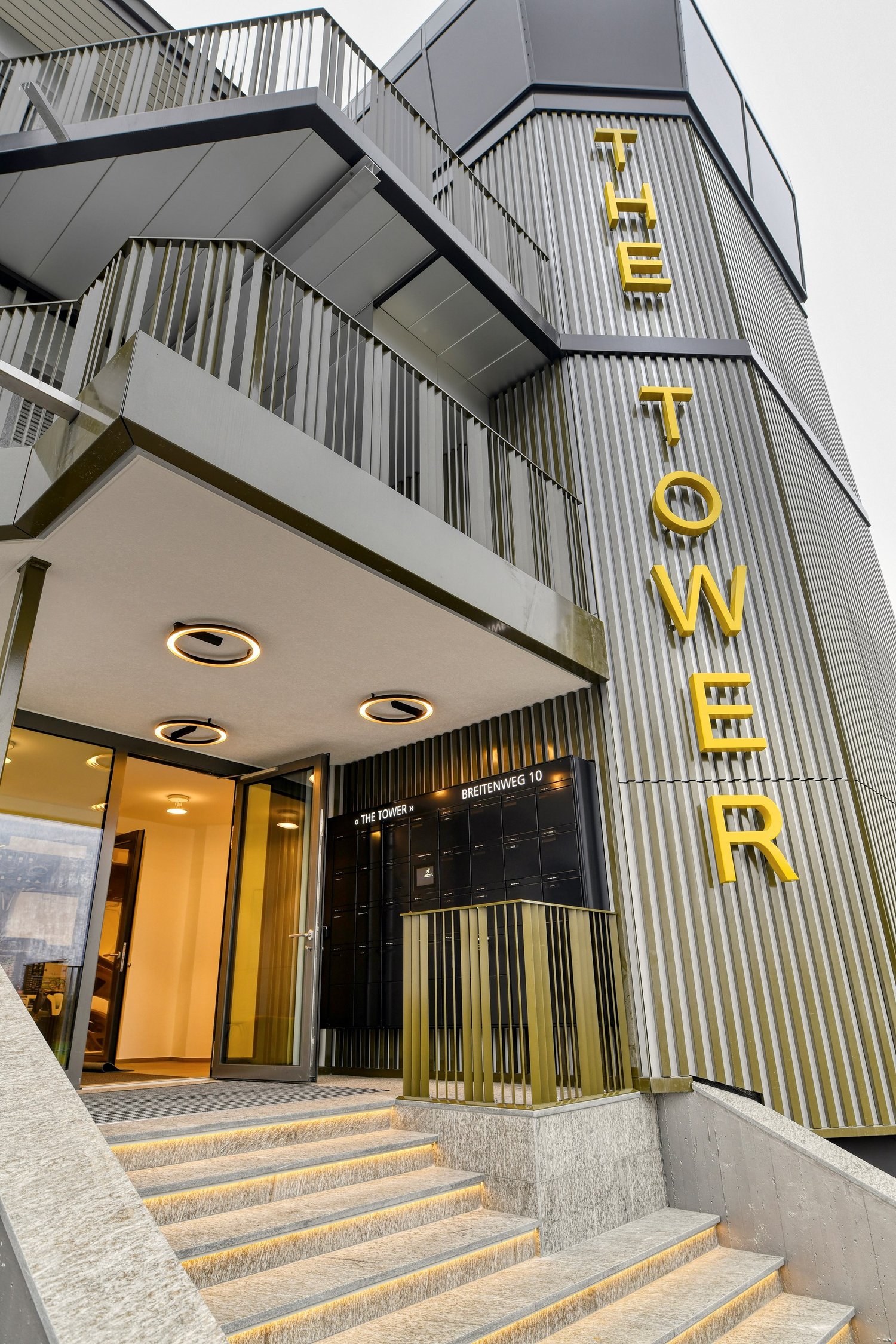 A modern multi-story building with steel exterior, glass doors, yellow branding, and a staircase leading to the entrance.