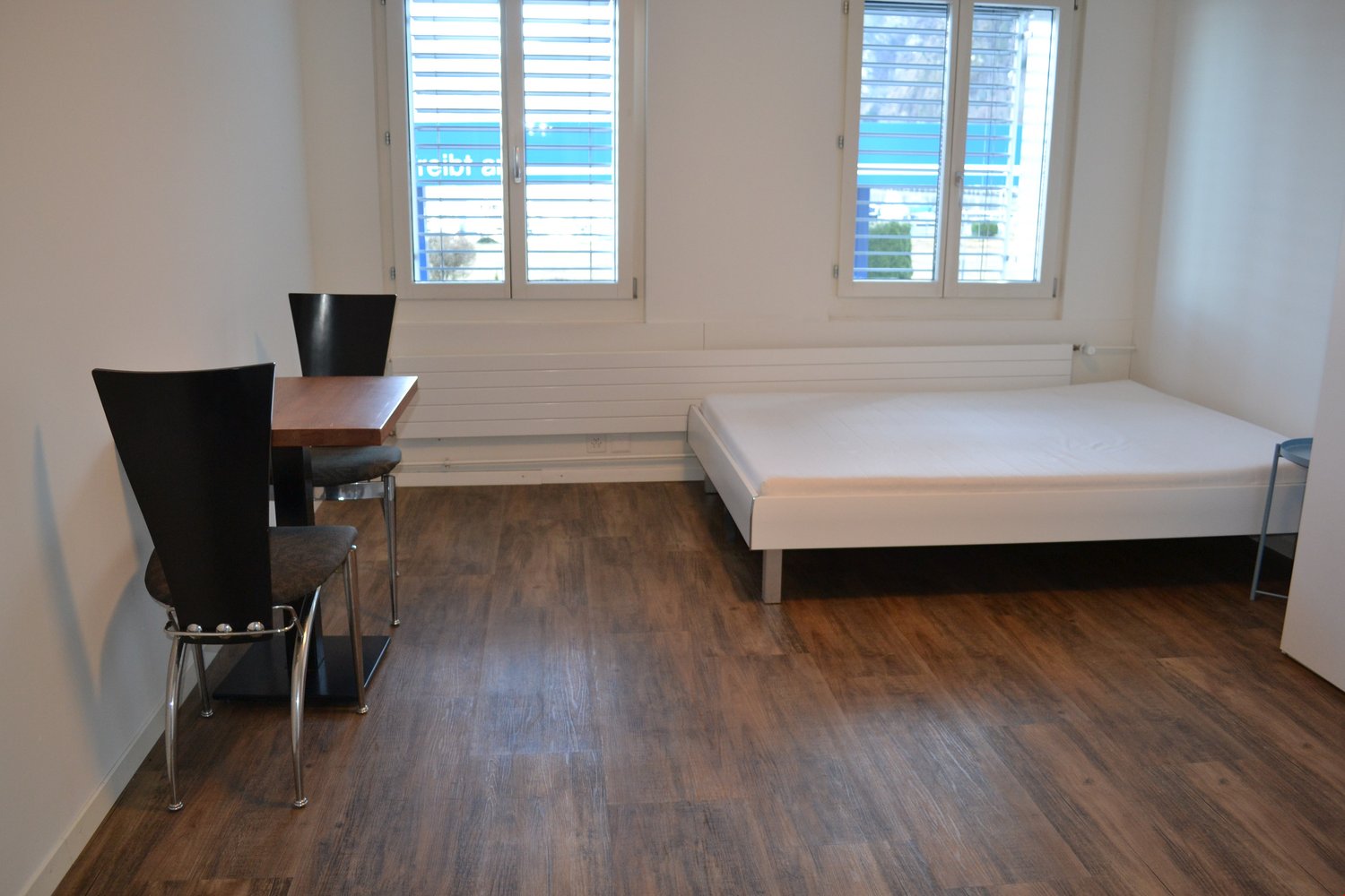 spacious room with wooden floor, bed, two chairs, wooden table, two windows, white walls