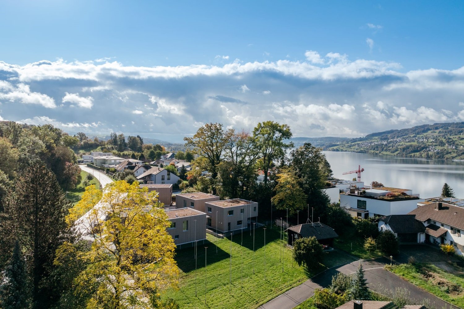 The image shows a scenic view of a lakeside town surrounded by lush greenery and autumn foliage. The town features a mix of residential buildings, some with red roofs, nestled among the trees. A body of water, likely a lake, can be seen in the distance, w
