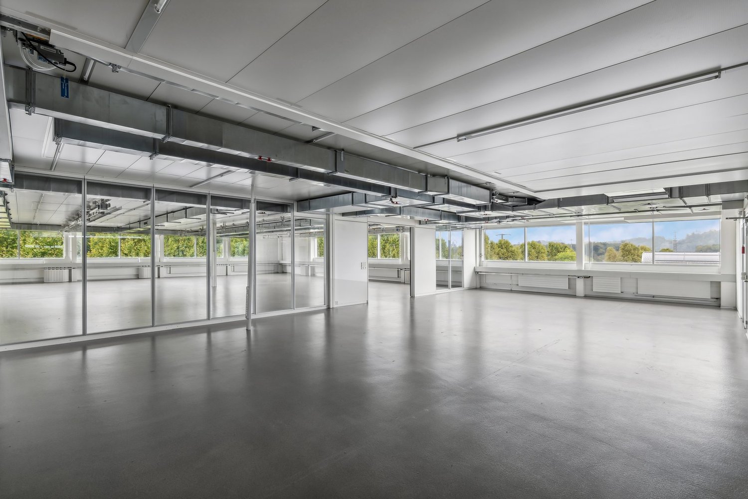 Large open office space with high ceilings, concrete floors, and floor-to-ceiling windows providing natural light and views of the surrounding greenery. The space has a modern, minimalist design with exposed ceiling beams and ductwork.