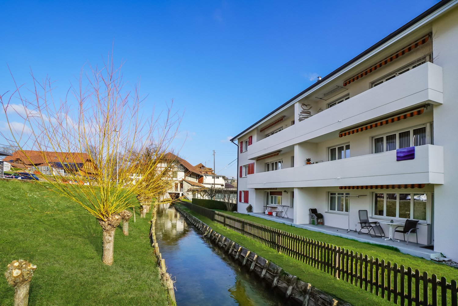 3 story white building, red shutters, balconies on each floor, small canal in front