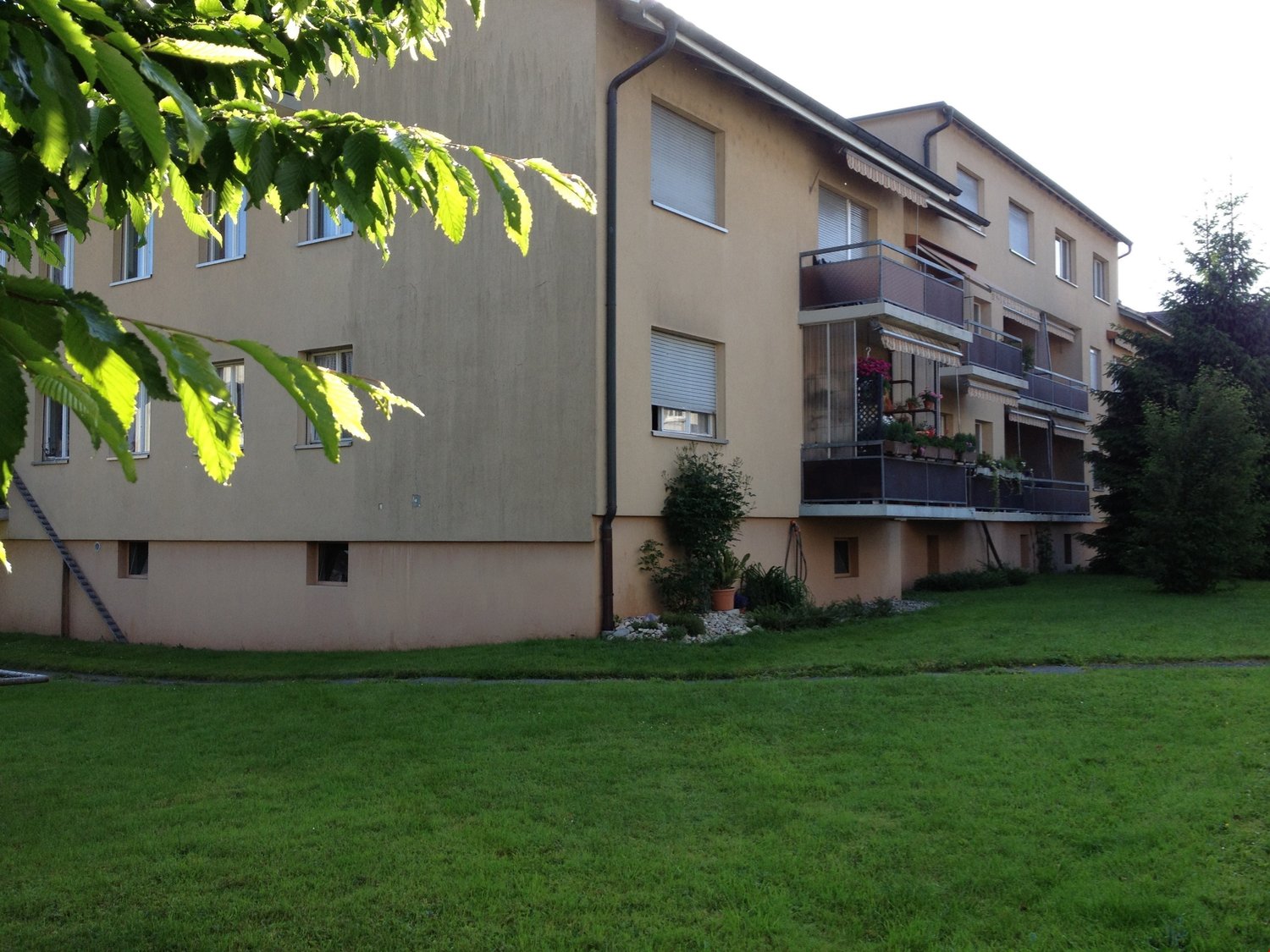 Apartment building, beige walls, three floors, multiple balconies, green lawn, trees, plants