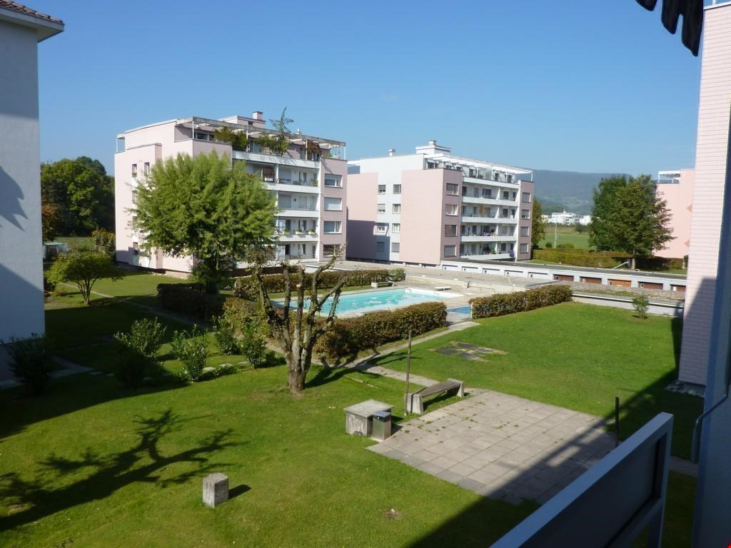 Swimming pool, green lawn, trees, bushes, benches, buildings in the background