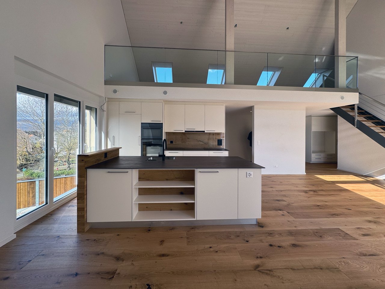 Open kitchen with central island and modern appliances, large windows, wooden floor, balcony, and a high ceiling