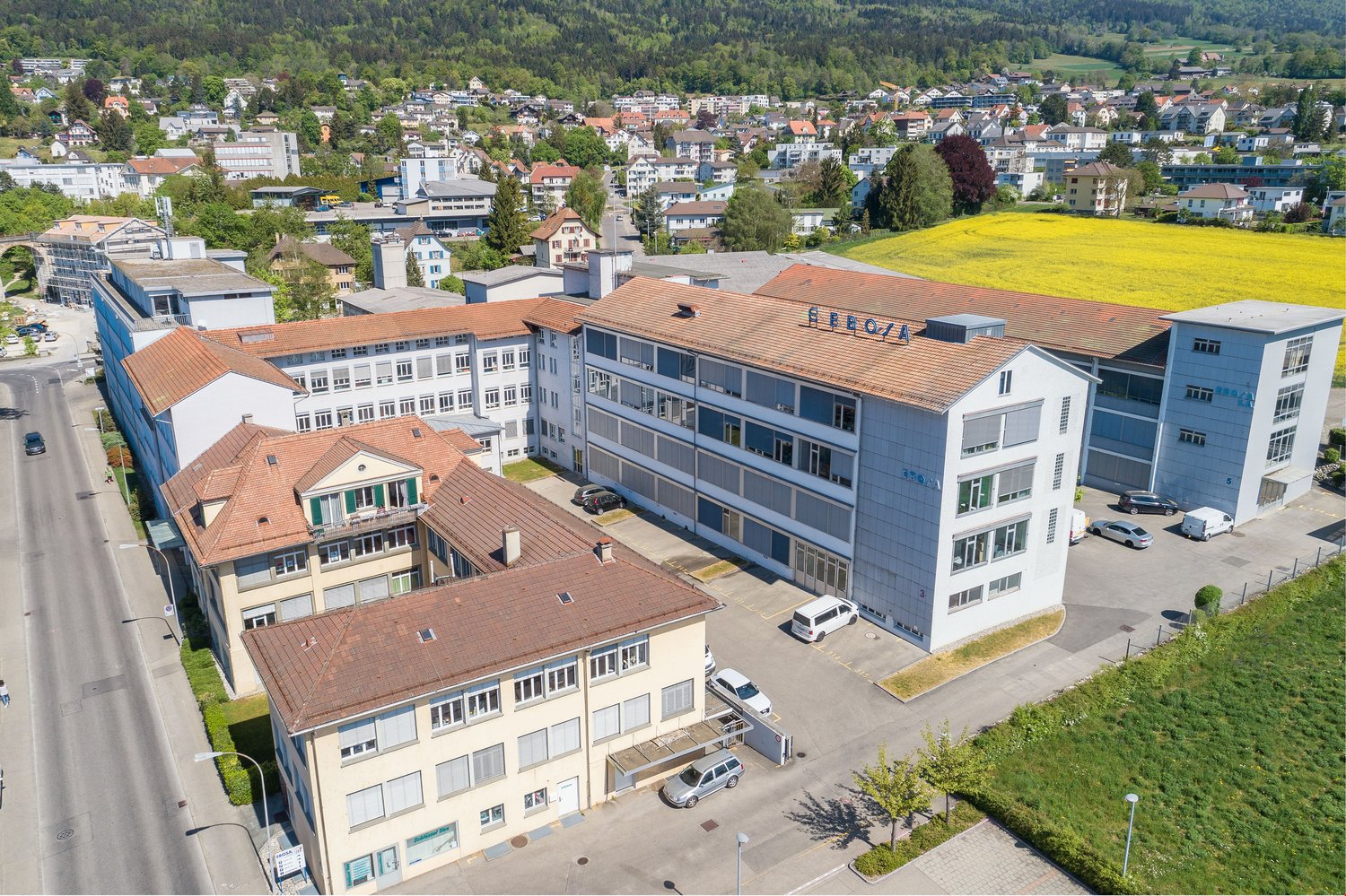 Multi-story commercial building with a large sign that says 'Erco/a', surrounded by other buildings and a grassy field in a hilly, forested area