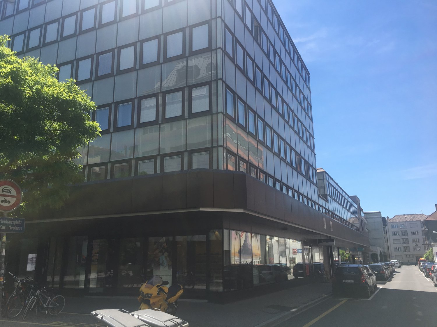 A multi-story glass and steel office building with a modern, sleek design. The building has a rectangular shape with rows of windows covering the facade. There are trees and other buildings visible in the surrounding area, indicating an urban setting.